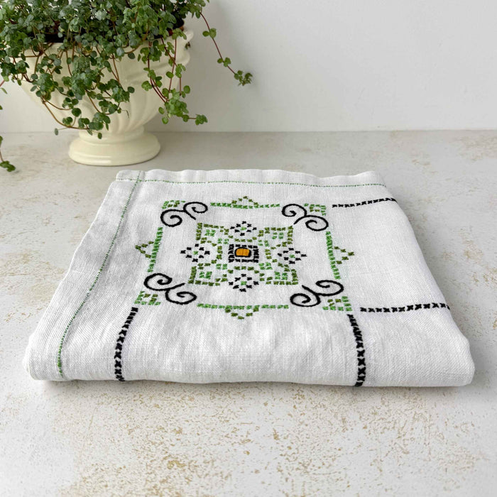 Low angle folded view of ecru embroidered tablecloth on a light surface, showing one geometric medallion in green, navy, and orange beside a potted plant.