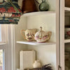 Hand-painted Lynn Hollyn sugar bowl and creamer with folk art style florals displayed on a white corner shelf beside a window, with vintage ceramics and dried lavender.
