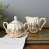 Floral sugar bowl and creamer displayed on a gold-toned tray with embroidered white linen and greenery.