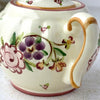 Close-up of sugar bowl showing loosely painted purple and pink flowers, green leaves, and golden brown handle decoration.