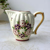 Hand-painted ceramic creamer with ribbed body, curved spout, golden brown handle, and stylized purple and pink floral bouquet.