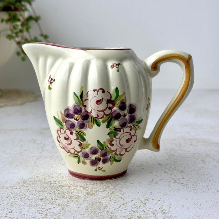 Hand-painted ceramic creamer with ribbed body, curved spout, golden brown handle, and stylized purple and pink floral bouquet.