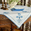 Off-white cross-stitch table topper draped over a dark wood table, with blue embroidered deer, trees and geometric motifs, finished with blue crocheted edging and styled with brass candlesticks.
