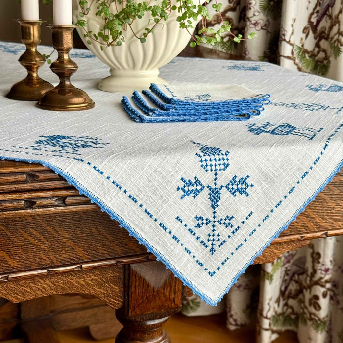 Off-white cross-stitch table topper draped over a dark wood table, with blue embroidered deer, trees and geometric motifs, finished with blue crocheted edging and styled with brass candlesticks.
