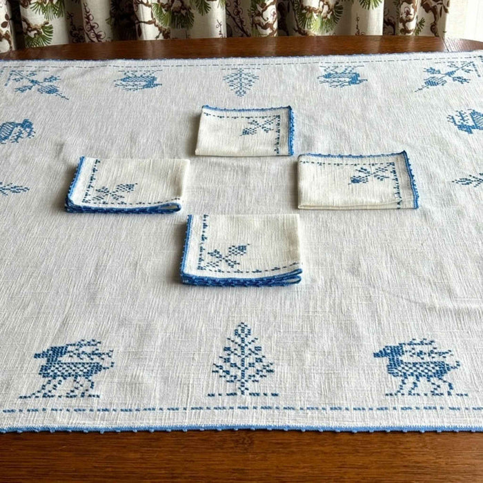 Blue and off-white embroidered table topper displayed on a dark wood table with matching napkins, showing the coordinated cross-stitch designs and blue crocheted borders.