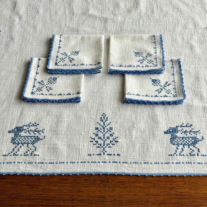 Close view of the off-white table topper draped over a dark wood table, showing stylized blue deer, trees, thistle-like motifs and crocheted edging.