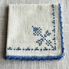 Folded off-white embroidered napkin showing blue cross-stitch decoration and a narrow blue crocheted edge.
