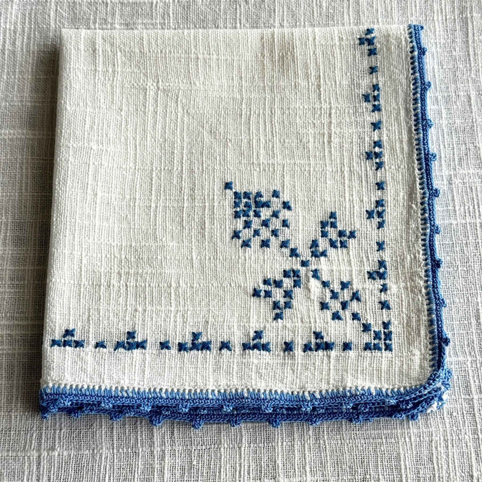 Folded off-white embroidered napkin showing blue cross-stitch decoration and a narrow blue crocheted edge.