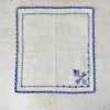Matching off-white napkin laid flat, showing its blue cross-stitch motif and crocheted edging.