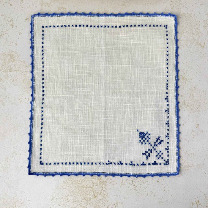 Matching off-white napkin laid flat, showing its blue cross-stitch motif and crocheted edging.