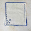 A second matching off-white napkin laid flat, showing the mirror-image arrangement of blue cross-stitch motifs and the blue crocheted border.
