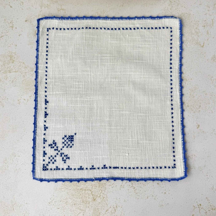 A second matching off-white napkin laid flat, showing the mirror-image arrangement of blue cross-stitch motifs and the blue crocheted border.