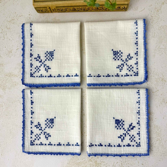 Matching embroidered napkins folded together, showing their off-white woven fabric, blue embroidery and coordinating crocheted edges.