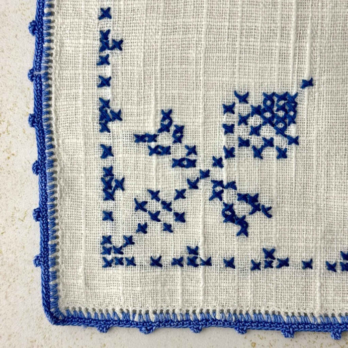 Close-up of the front of napkin showing blue cross-stitched thistle on textured off-white fabric.