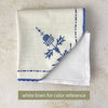 Off-white embroidered table linen placed against a white napkin for color comparison, showing its warmer off-white tone and blue cross-stitch decoration.