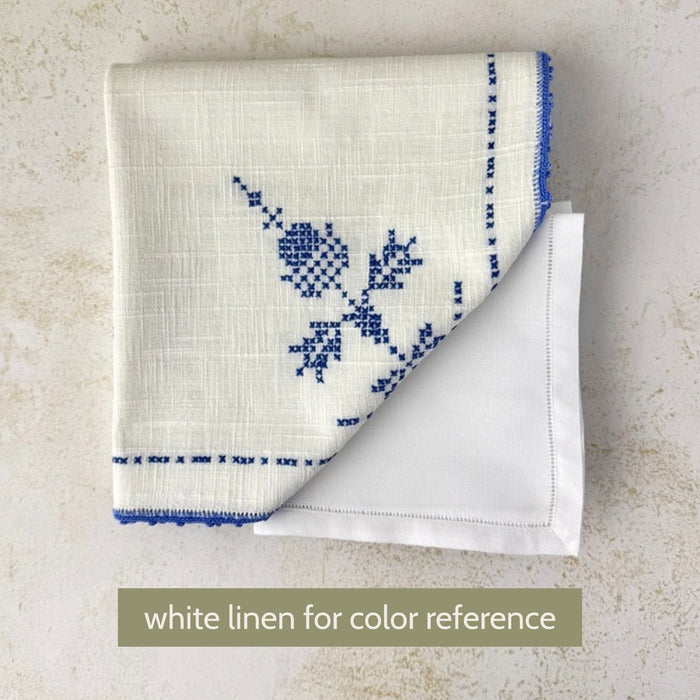 Off-white embroidered table linen placed against a white napkin for color comparison, showing its warmer off-white tone and blue cross-stitch decoration.