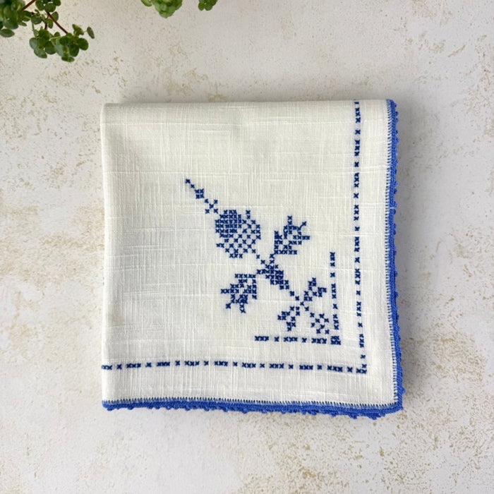 Overhead view of the folded off-white table topper showing several blue embroidered motifs, the textured woven fabric and crocheted edging.