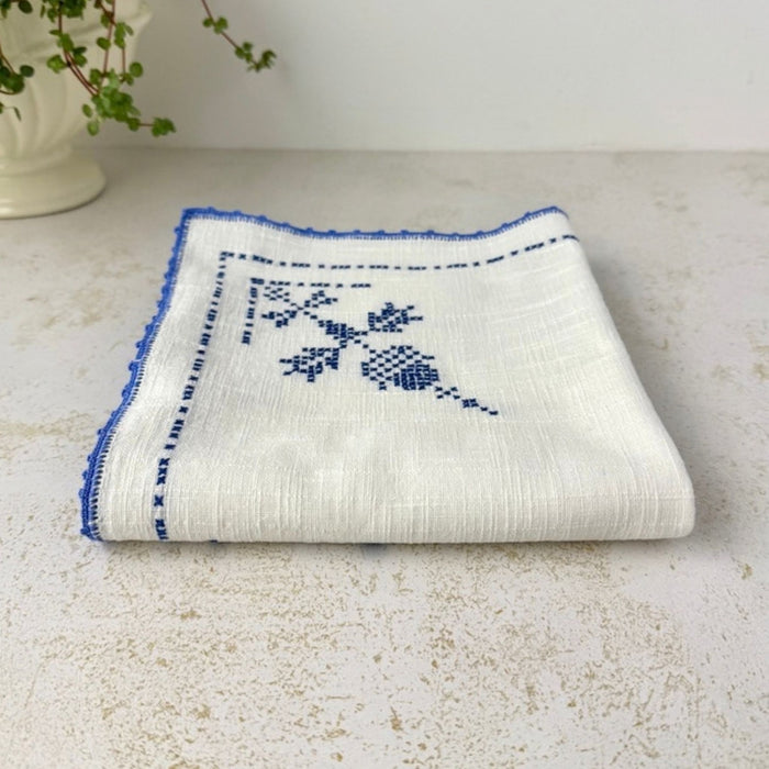 Low-angle view of the folded table topper showing the fabric's substantial weave, layered folds, blue cross-stitch decoration and crocheted border.
