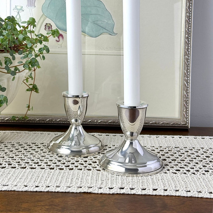Pair of Duchin sterling silver candlesticks holding white taper candles, styled on a crocheted table runner in front of framed botanical artwork.