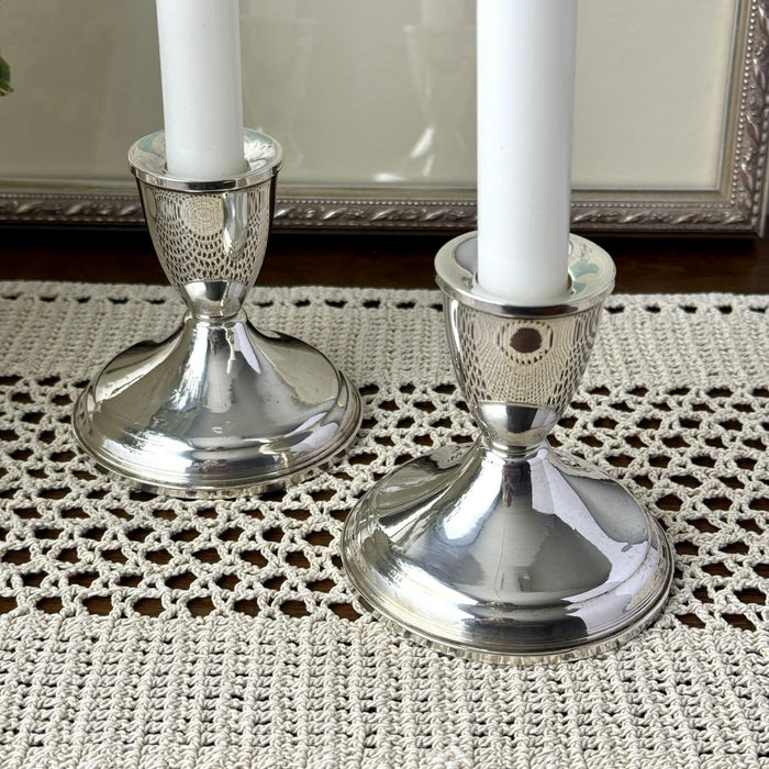 Close-up of two polished sterling silver candlesticks with white taper candles, showing their gently tapered stems, rounded bases, and reflective surfaces.