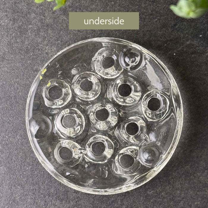 Underside of a clear glass flower frog showing eleven openings passing through the thick glass body and 3 feet.