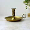 Vintage brass chamberstick with a wide round drip pan, tall central candle cup, and finger loop handle, shown against a light background.