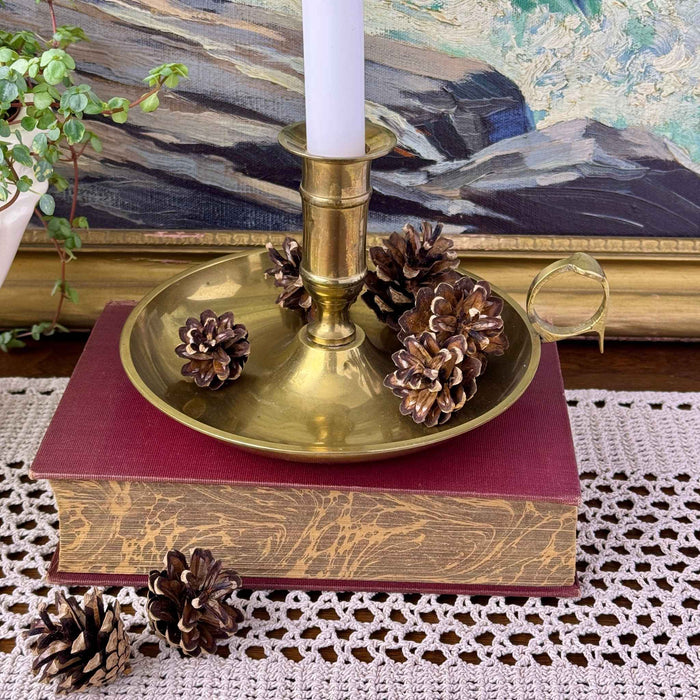 Brass chamberstick holding a white taper candle, styled with pinecones on a burgundy book over a crocheted table runner.