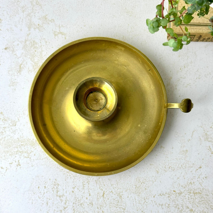 Overhead view of a brass chamberstick showing its wide circular drip pan, central candle cup, and side handle.