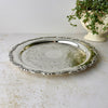 Round silverplate serving tray with an ornate scalloped floral border and intricate scrolling decoration across the center, photographed from above.