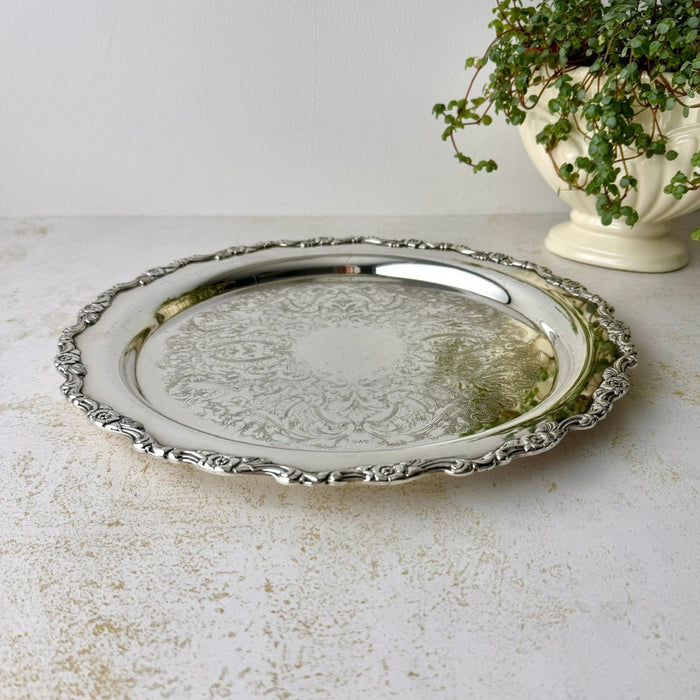 Round silverplate serving tray with an ornate scalloped floral border and intricate scrolling decoration across the center, photographed from above.