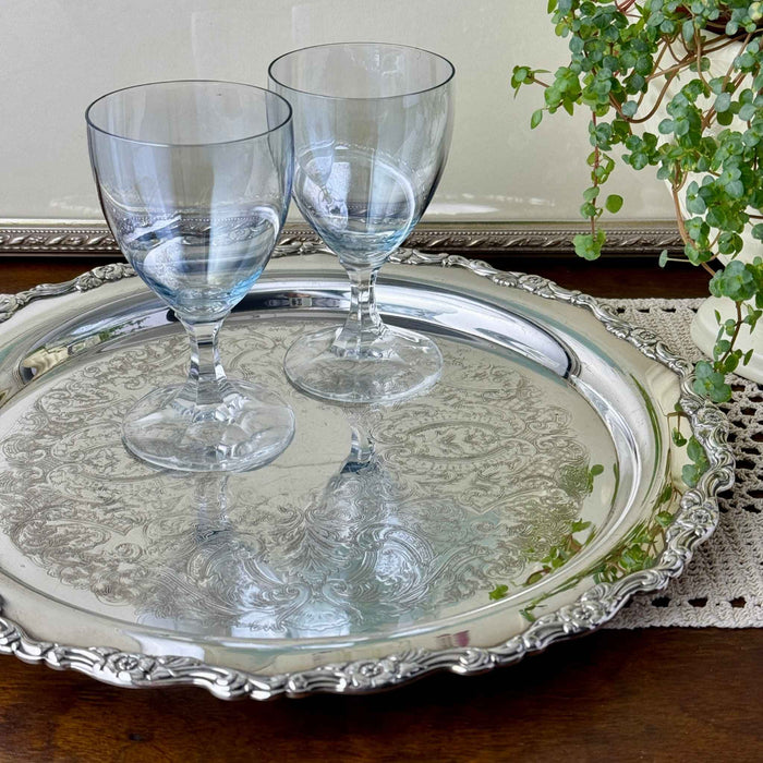 Ornate silverplate serving tray holding two clear stemmed glasses on a wooden table, with a trailing green plant and crocheted linen nearby.