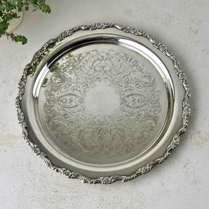 Round silverplate serving tray with an ornate scalloped floral border and intricate scrolling decoration across the center, photographed from above.