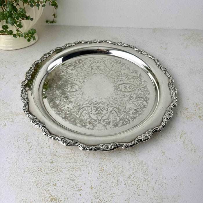 Round silverplate tray photographed at an angle, showing its wide raised rim, scalloped floral border, and decorated center.