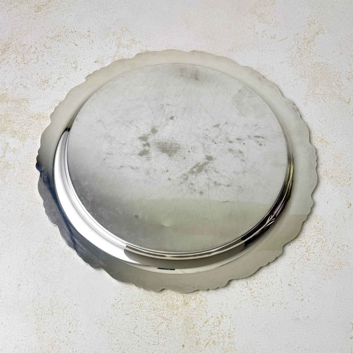 Underside of the round silverplate tray showing its circular base, numerous surface scratches, scattered dark spots, and scalloped outer edge.