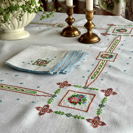 Off-white cross-stitch tablecloth with colorful geometric floral embroidery, styled on a dark wood table with four folded matching napkins, brass candlesticks, and a cream planter.