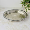 Oval silverplate gallery tray with a pierced railing, patterned raised rim, and elaborate floral scrollwork across the base, photographed beside a trailing green plant.