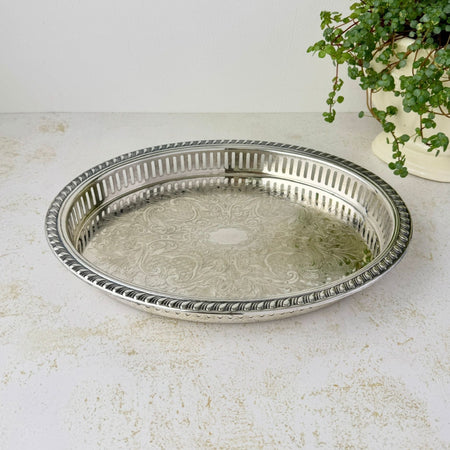 Oval silverplate gallery tray with a pierced railing, patterned raised rim, and elaborate floral scrollwork across the base, photographed beside a trailing green plant.