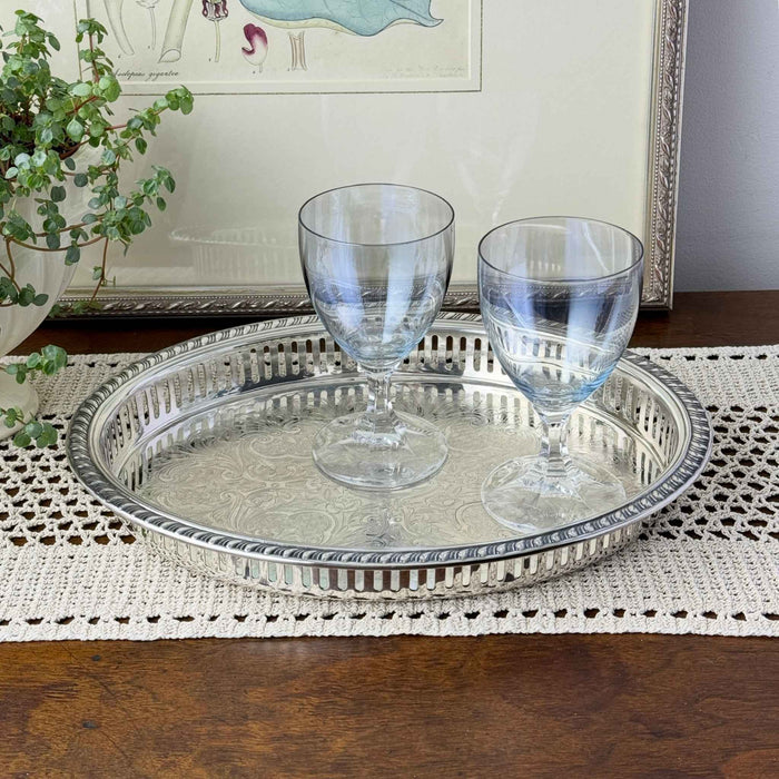 Oval silverplate gallery tray holding two clear stemmed glasses on a crocheted runner, with a framed botanical print and potted plant behind it.