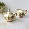 Matching hand-painted ceramic sugar bowl and creamer with ribbed cream-colored bodies, purple and pink stylized florals, and golden brown edged handles.