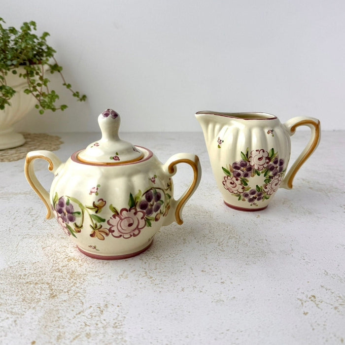 Matching hand-painted ceramic sugar bowl and creamer with ribbed cream-colored bodies, purple and pink stylized florals, and golden brown edged handles.