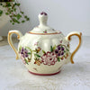 Front view of lidded sugar bowl with two curved handles, ribbed body, purple and pink floral decoration, and painted lid.