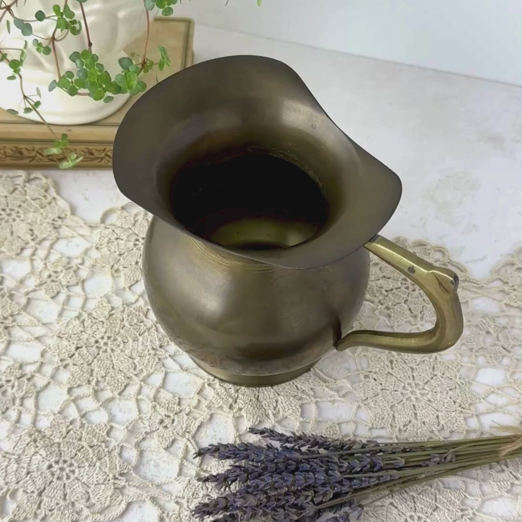 Close view of a vintage brass pitcher with aged patina, panning from the open rim and interior down to reveal the rounded body, curved handle, and full profile