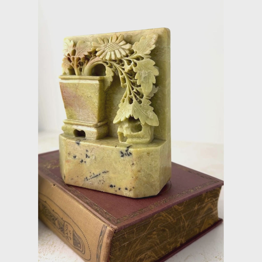 Close-up view of carved soapstone bookends displayed with books panning from one bookend to the other showing dimensional potted plants, flowers and natural stone variation