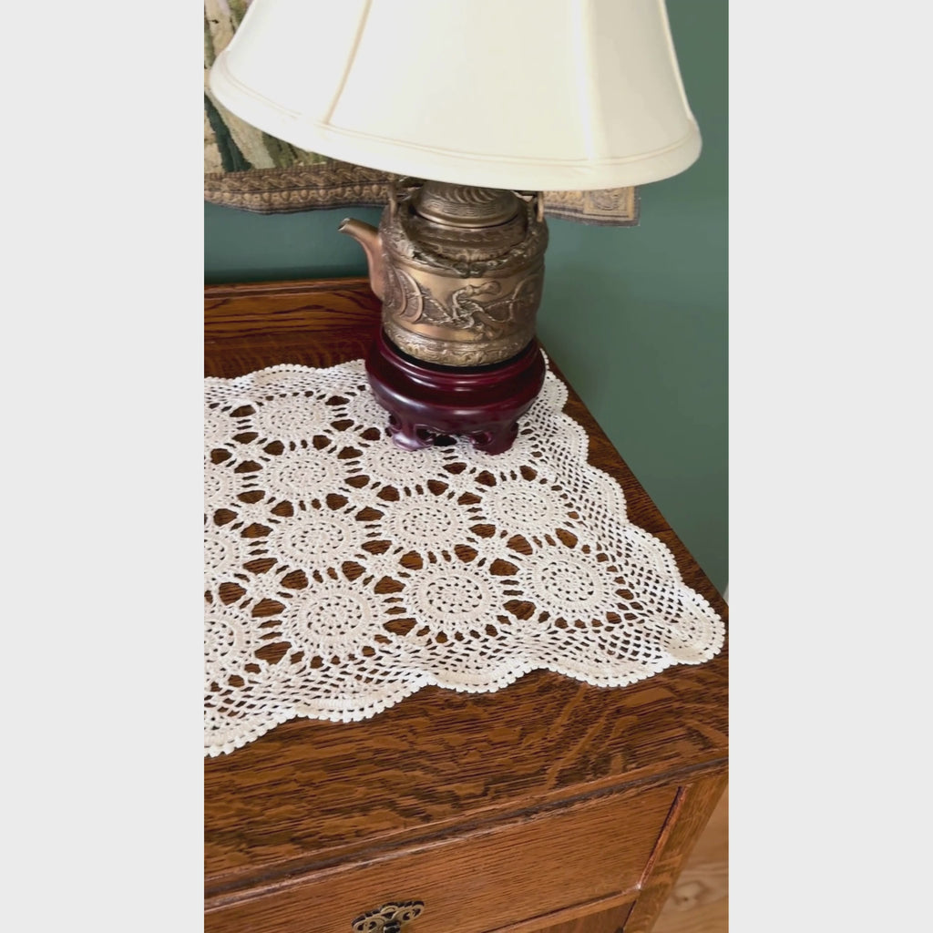 Video of ecru crochet table runner styled across a dark wood table with brass candlesticks, white candles, and a white planter with greenery.