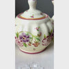 Close-up video of matching hand-painted ceramic sugar bowl and creamer with ribbed cream-colored bodies, purple and pink stylized florals, and golden brown edged handles.