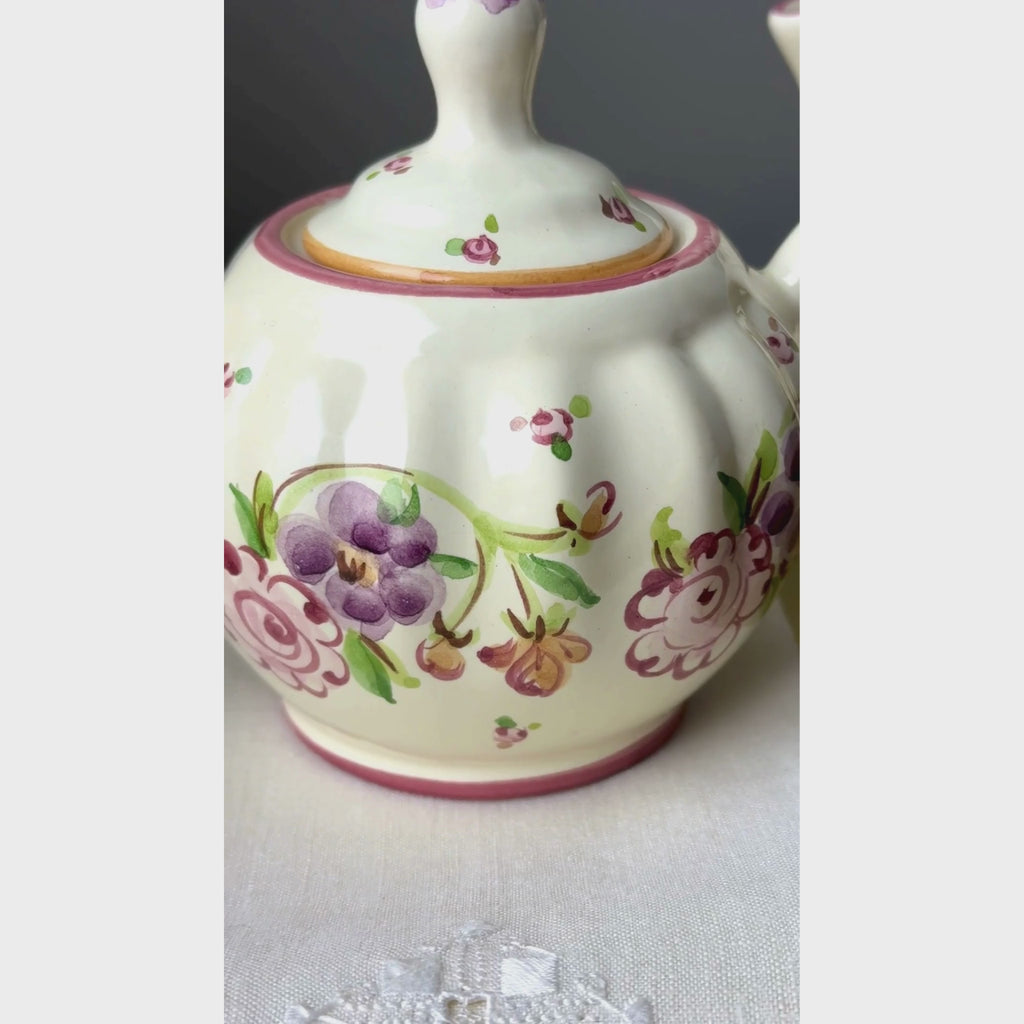 Close-up video of matching hand-painted ceramic sugar bowl and creamer with ribbed cream-colored bodies, purple and pink stylized florals, and golden brown edged handles.