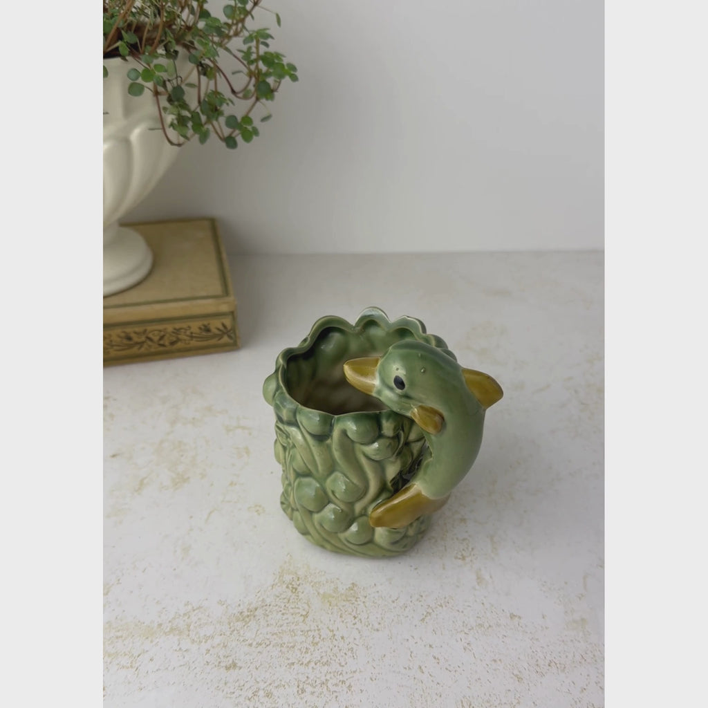 Vintage dolphin and waves planter shown on a neutral backdrop, picked up and turned by hand to show its surface and interior