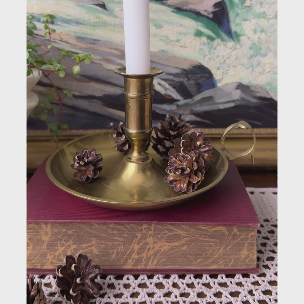 Video of vintage brass chamberstick holding a white taper candle, styled with pinecones on a burgundy book.
