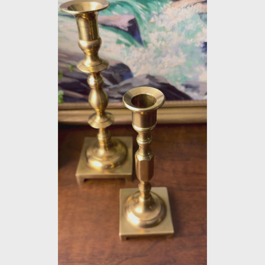 Video of mismatched pair of vintage solid brass candlesticks with turned stems, square footed bases, and staggered heights.