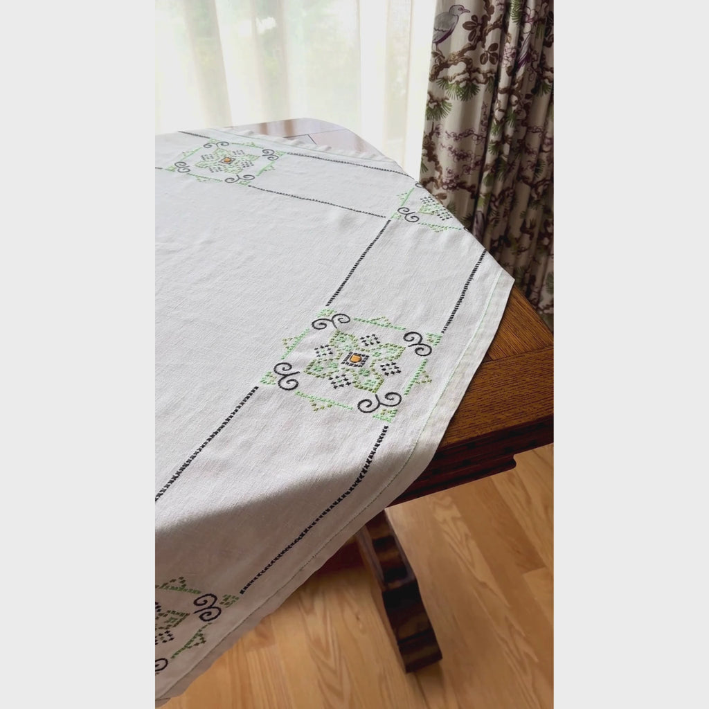 Video of ecru square embroidered tablecloth draped diagonally over a dark wood table, showing navy and green geometric medallion borders with small orange centers.